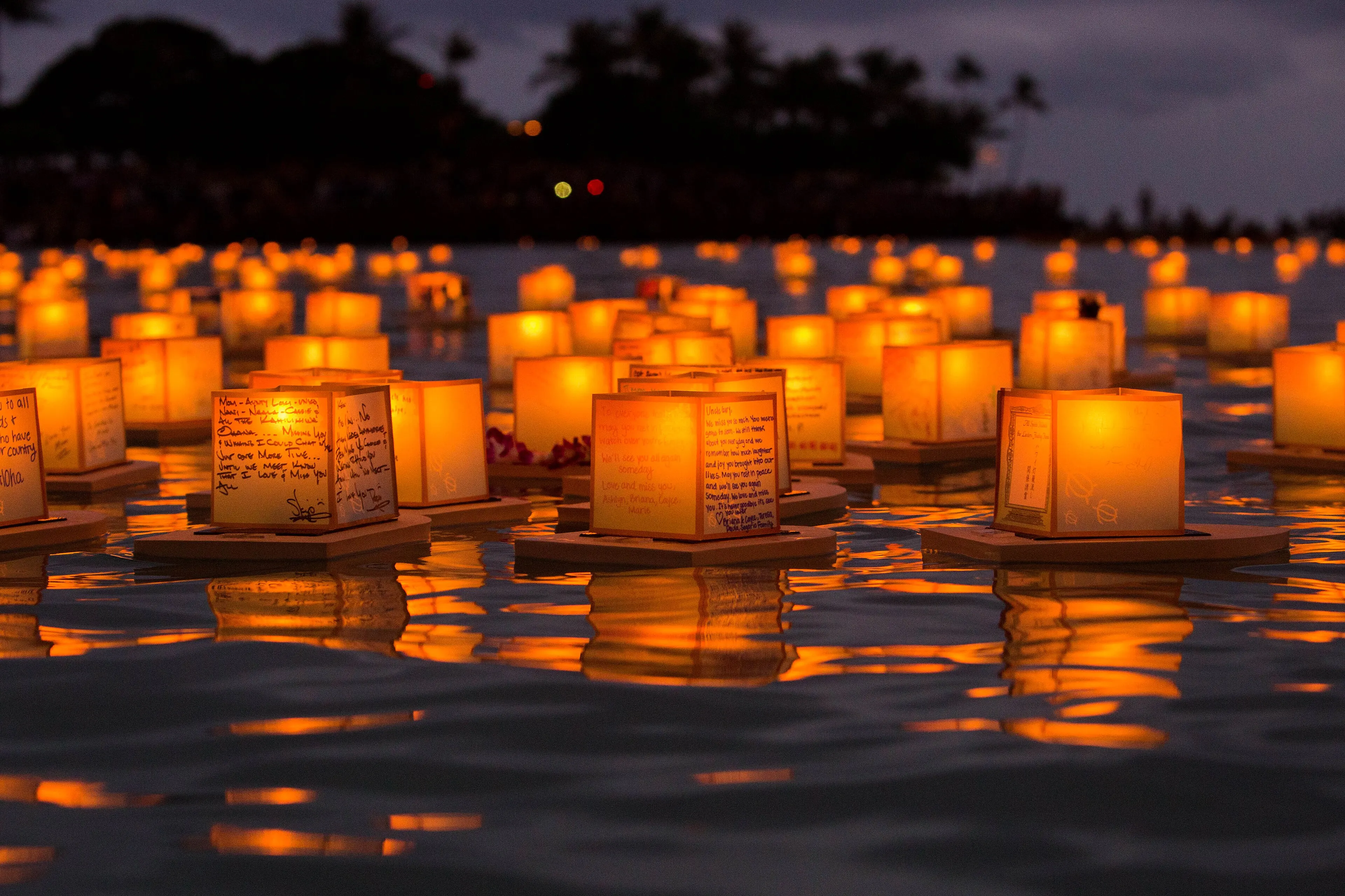 Glowing paper lanterns with handwritten messages floating on the water at dusk during the Lantern Floating Hawaiʻi ceremony
