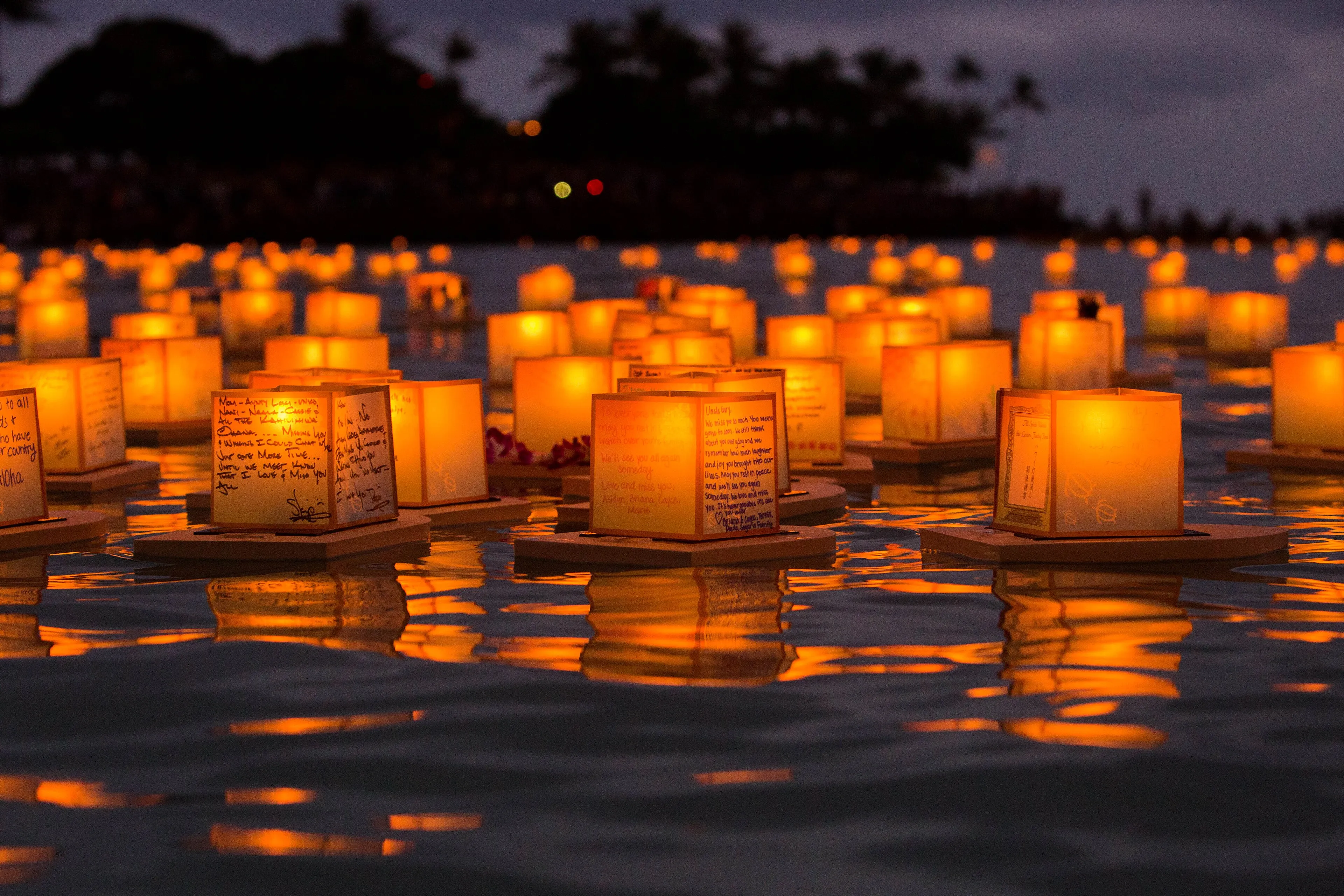 Glowing paper lanterns with handwritten messages floating on the water at dusk during the Lantern Floating Hawaiʻi ceremony