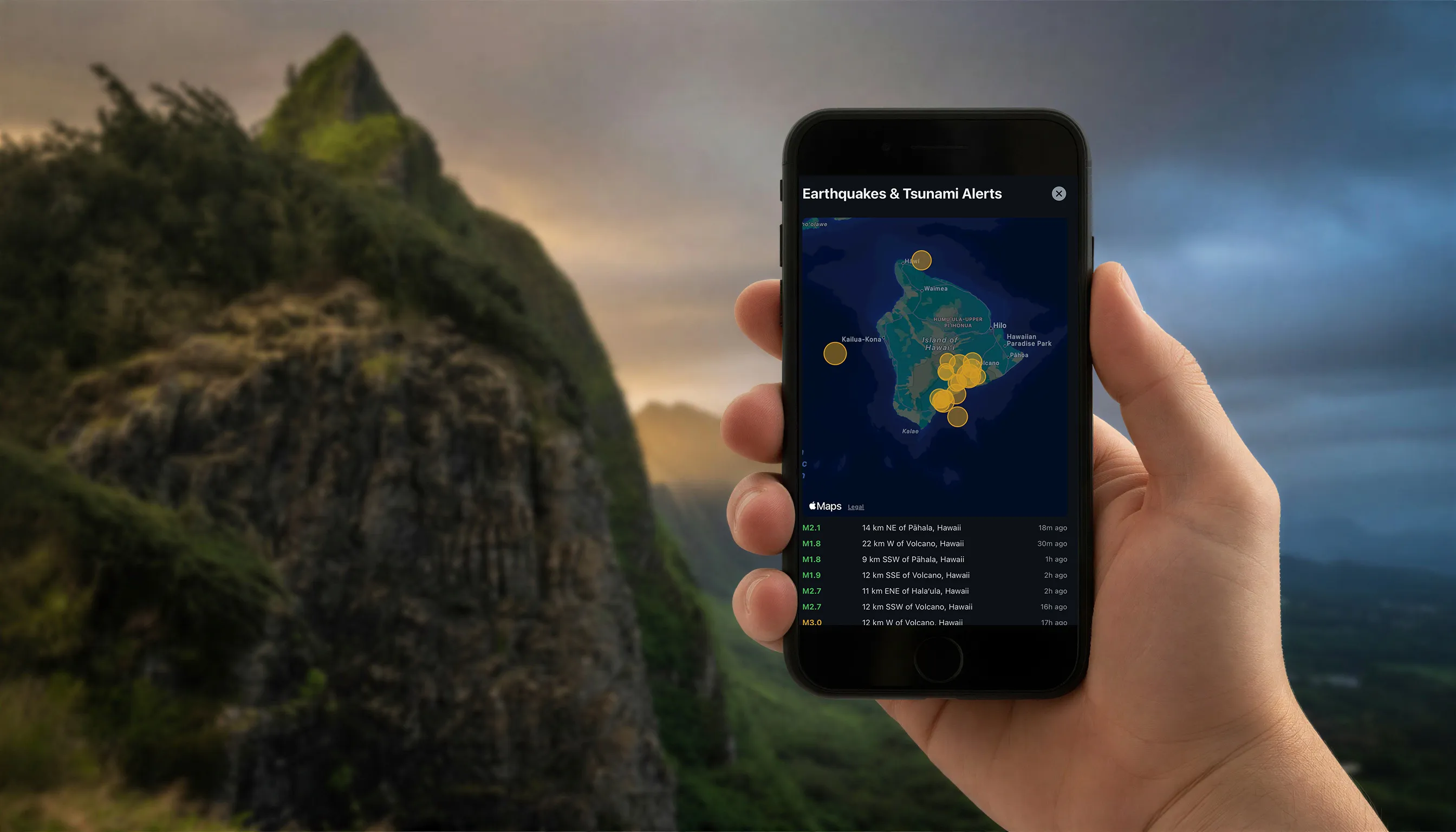 Tapiki StormWatch on an iPhone, held up against the Koʻolau mountains under storm clouds, showing the Earthquakes & Tsunami Alerts map of Hawaiʻi Island